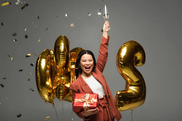 Excited Asian Woman Holding Present Champagne Balloons Confetti Grey Background — Stock Photo, Image