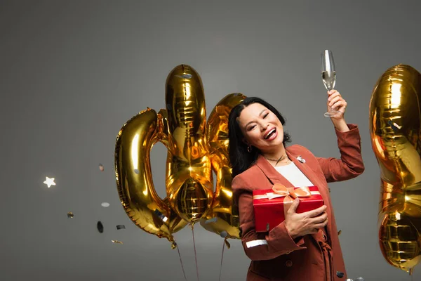 带着礼物和香槟酒的快乐的亚洲女人 在气球和灰色背景的圆饼旁边看着相机 — 图库照片