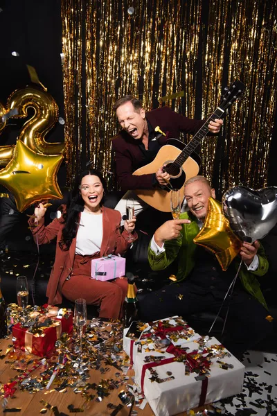 黒を背景に新年を祝うシャンパン アコースティックギター 風船と肯定的な異人種間の友人 — ストック写真