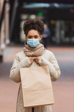 Esmer, Afro-Amerikalı, tıbbi maskeli bir kadın alışveriş merkezinde alışveriş çantası tutuyor. 