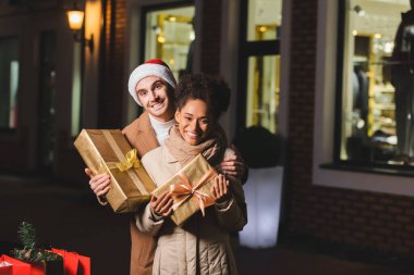 Noel Baba şapkalı mutlu adam ve Afrikalı Amerikalı kız arkadaşı Noel hediyesi kutularını tutuyorlar.