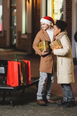 Noel Baba şapkalı mutlu adam ve Afro-Amerikalı kız arkadaşı Noel hediyesi kutularını tutarken alışveriş çantalarına bakıyor.