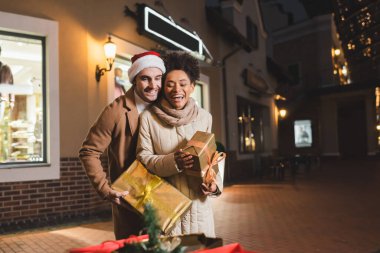 Noel Baba şapkalı mutlu adam ve Afrikalı Amerikalı kız arkadaşı elinde Noel hediyesi kutuları tutarken bulanık alışveriş çantalarına bakıyor.