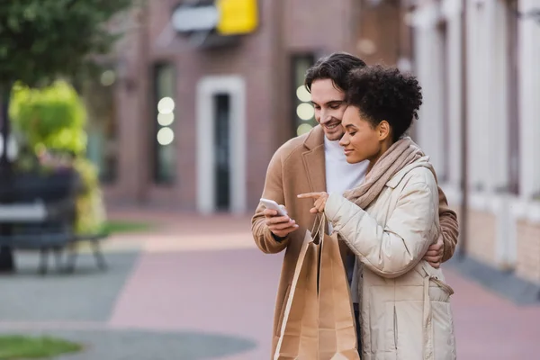 若いアフリカ系アメリカ人の女性が買い物袋を持っている彼氏の近くのスマートフォンを指して — ストック写真