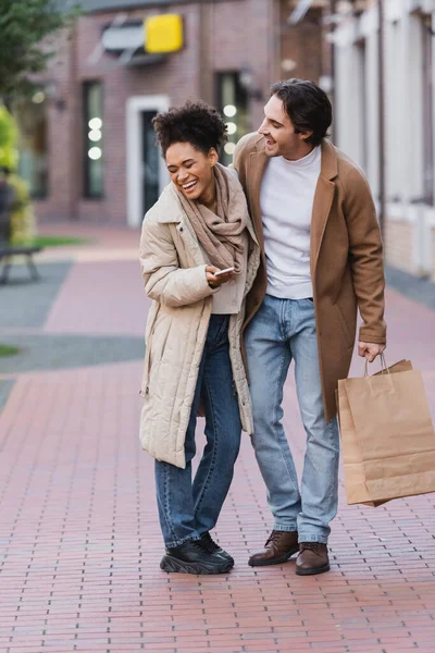 幸せなアフリカ系アメリカ人女性スマートフォンは幸せなボーイフレンドの近くで笑ってショッピングバッグを保持 — ストック写真