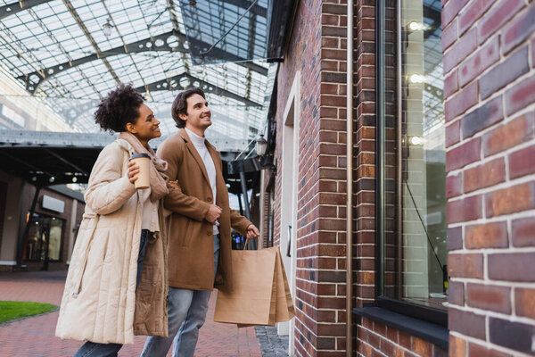 happy african american woman with paper cup standing with boyfriend holding shopping bags near window in mall
