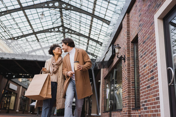 happy african american woman with shopping bags walking near boyfriend holding paper cup