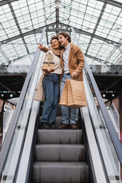 african american woman holding coffee to go near man with shopping bags pointing away on escalator in mall