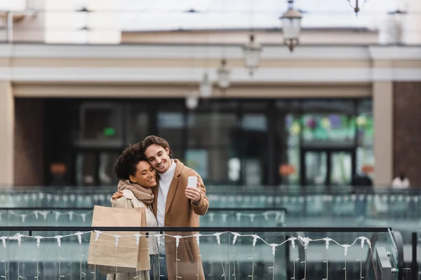 Glada Multietniskt Par Med Papperskoppar Och Shoppingväskor Som Tar Selfie — Stockfoto