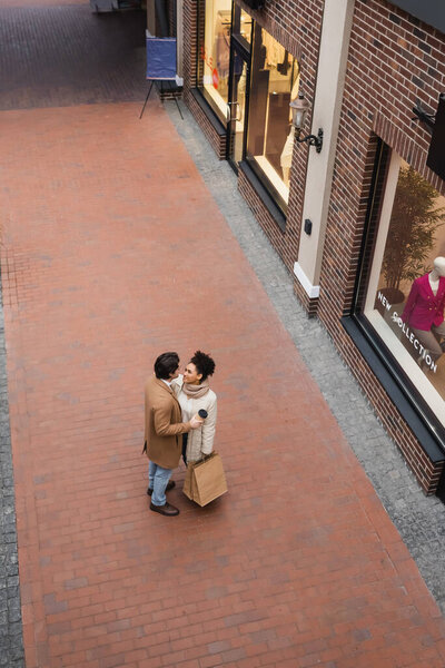 high angle view of man holding paper cup near pleased african american girlfriend with purchases in mall 