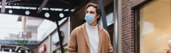 医療用マスクとベージュのコートを着た若者がショッピングセンターバナーを歩いていて — ストック写真