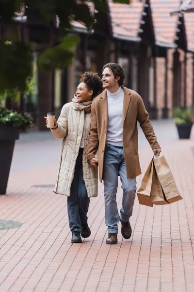 Alegre Pareja Multiétnica Abrigos Tomados Mano Mientras Camina Con Compras —  Fotos de Stock