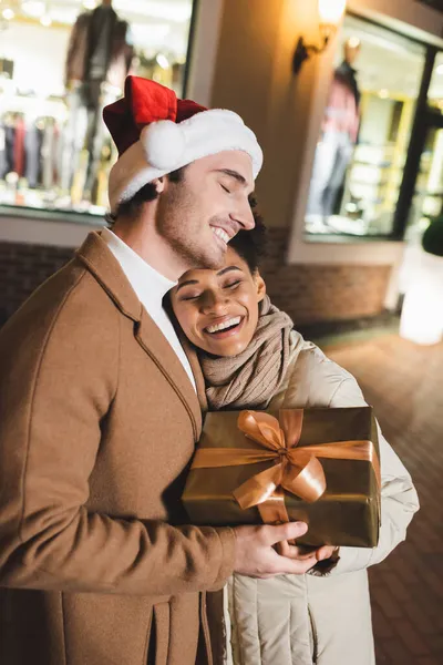 Mutlu Afro-Amerikalı kadın ve Noel Baba şapkalı memnun adam akşamları hediye paketi taşıyor. 
