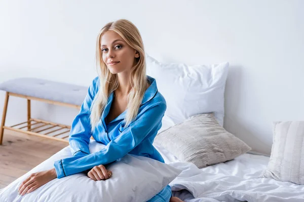ベッドに座りながら枕を持つ陽気な若い女性 — ストック写真