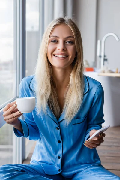 Feliz Joven Mujer Sosteniendo Teléfono Celular Taza Café Cerca Ventana — Foto de Stock