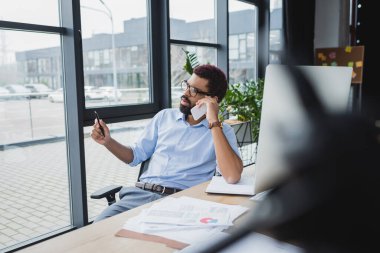 Afrika kökenli Amerikalı işadamı, ofiste bilgisayar ve kağıt yakınlarında akıllı telefondan konuşuyor. 
