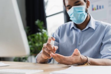 Bulanık Afro-Amerikan iş adamı tıbbi maskeli. Ofisteki bilgisayarın yanında el dezenfektanı sıkıyor. 