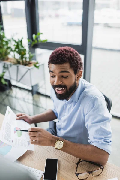 アフリカ系アメリカ人のビジネスマンがオフィスのスマートフォンやコンピュータの近くの紙を指して笑顔で — ストック写真