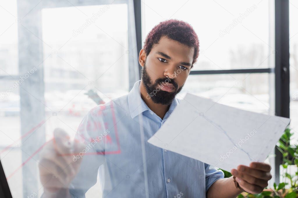 African american businessman looking at document and writing on blurred glass board in office 