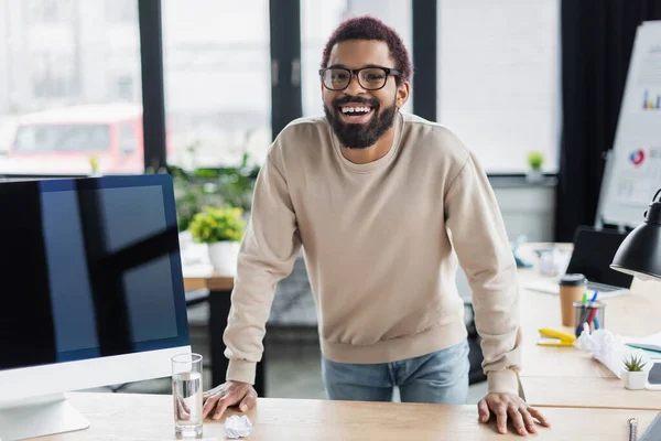 オフィスで水とコンピュータモニタの近くのカメラで微笑む陽気なアフリカ系アメリカ人ビジネスマン — ストック写真