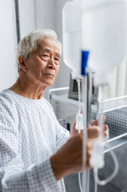 Asian patient in gown looking at blurred intravenous therapy in hospital ward clipart