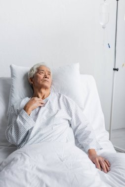 Sick asian man lying on bed near intravenous therapy in hospital ward  clipart