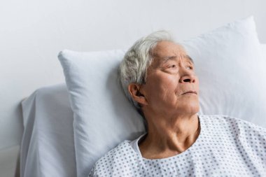 Klinikte yatağa uzanmış önlüklü yaşlı Asyalı hasta. 