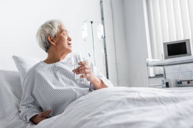Elderly asian patient holding glass of water on hospital bed  clipart