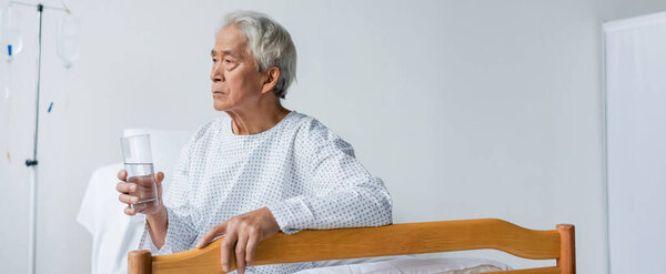 Старший азиатский пациент держит стакан воды на кровати в палате больницы, баннер 