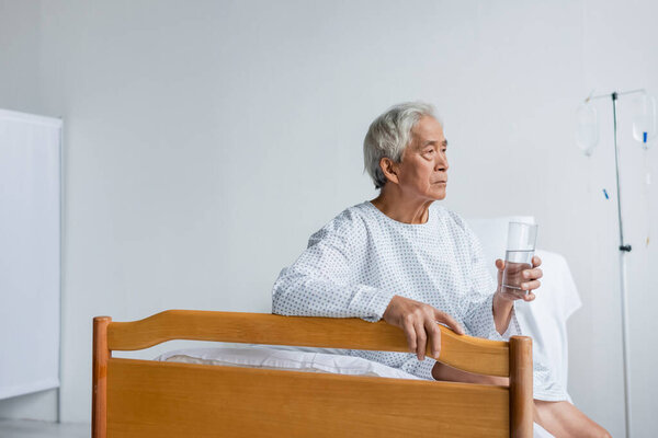 Пожилой азиатский пациент, держащий стакан воды на кровати в палате больницы 