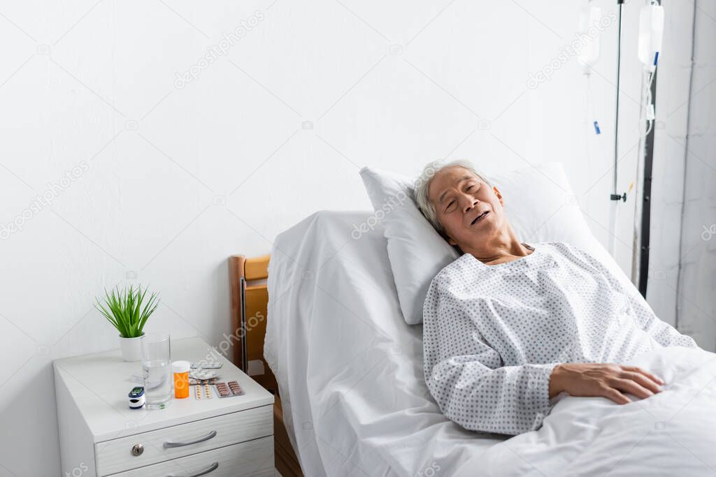 Ill asian patient lying on bed near pills and water in hospital ward 