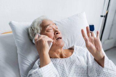 Sabahlık ve nabız ölçer takmış Asyalı bir hasta klinikteki yatakta akıllı telefonla konuşuyor. 