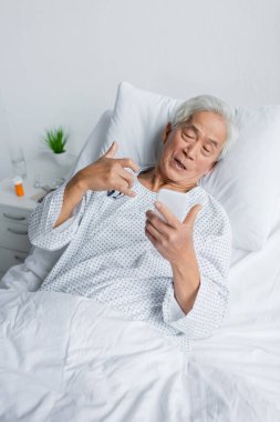Nabız oksimetresi olan yaşlı Asya patenti. Klinikte yatakta akıllı telefon kullanıyor. 