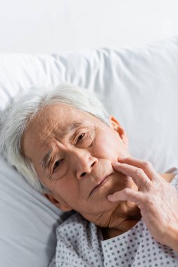 Yaşlı Asyalı hasta klinikte yatakta yatarken kameraya bakıyor. 