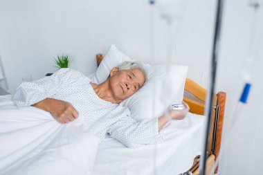 Senior asian man with pulse oximeter lying near blurred intravenous therapy on bed in hospital  clipart