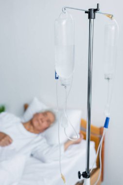 Intravenous therapy stand near blurred patient on bed in hospital  clipart