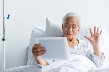 Burun kanülü olan kıdemli Asyalı hasta klinikteki dijital tablete video çağrısı yapıyor. 