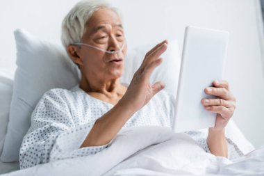 Hastane koğuşunda burun kanülü olan bulanık Asyalı bir hastanın elindeki dijital tablet. 