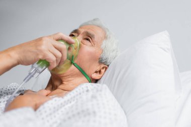 Yaşlı Asyalı hasta klinikte yatakta oksijen maskesi soluyor. 