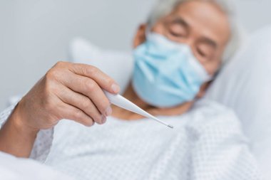 Tıbbi maskeli bulanık Asyalı hasta klinikteki yatakta termometre tutuyor. 