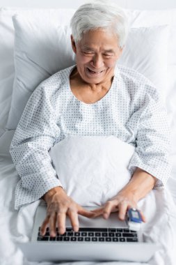 Nabız oksimetresi olan pozitif Asyalı hasta klinikteki yatakta bulanık dizüstü bilgisayar kullanıyor. 