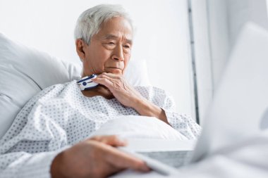 Nabız oksimetresi olan yaşlı Asyalı hasta hastane koğuşunda bulanık dizüstü bilgisayar tutuyor. 