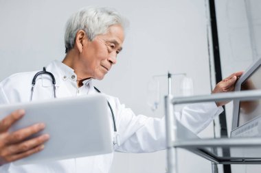 Klinikteki tıbbi ekipmanların yanında bulanık dijital tablet tutan yaşlı Asyalı doktor 
