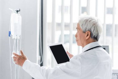 Dijital tabletli yaşlı Asyalı doktorun klinikteki damar içi terapi standına bakması. 