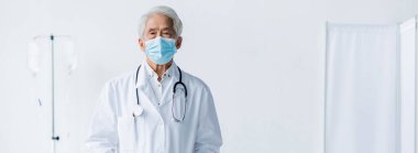Tıbbi maskeli ve beyaz önlüklü yaşlı Asyalı doktor klinikteki kameraya bakıyor. 