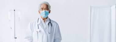 Elderly asian doctor in medical mask and white coat looking at camera in clinic, banner  clipart
