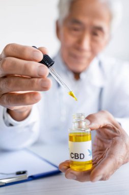 Cbd oil in hands of blurred asian doctor near clipboard in clinic clipart