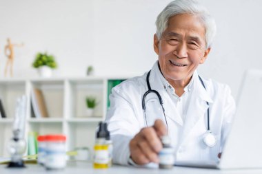 Gülümseyen Asyalı doktor klinikteki tıbbi esrar ve dizüstü bilgisayarın yakınındaki kameraya bakıyor. 