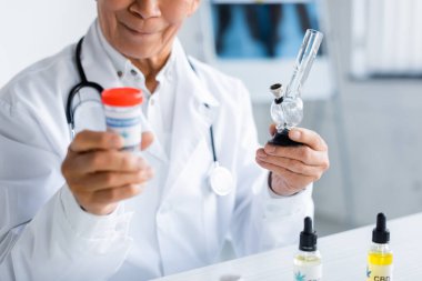 Klinikteki CBD yağının yanında elinde nargile ve tıbbi esrar tutan doktor görüntüsü. 