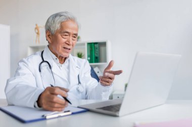 Yaşlı Asyalı doktor video görüşmesi sırasında bilgisayarı işaret ediyor ve klinikteki panoya yazı yazıyor. 